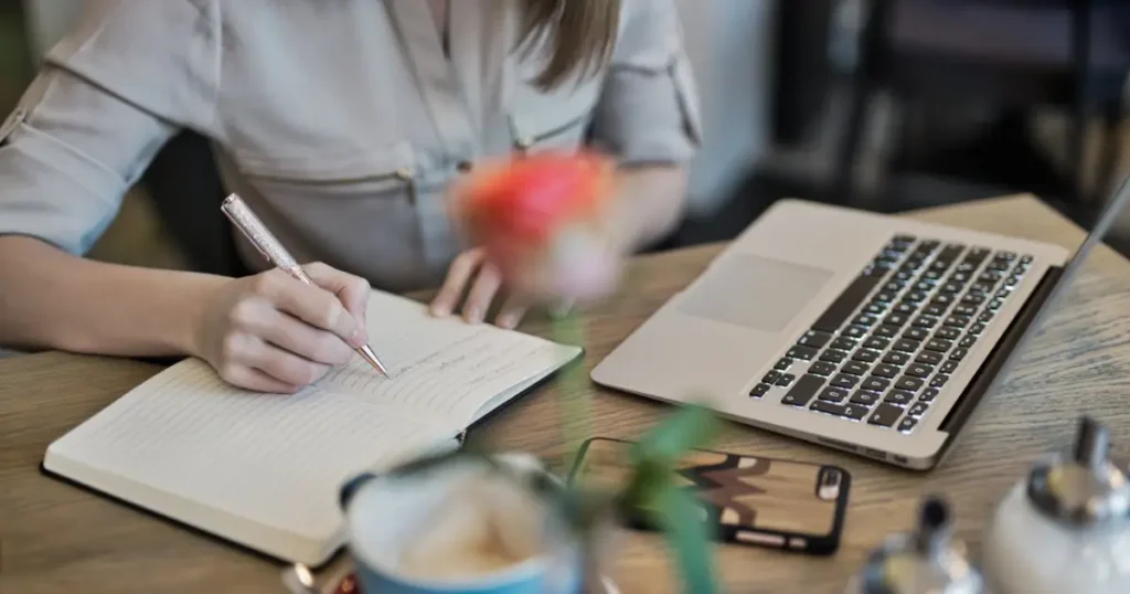 a person writing in a notebook next to a laptop